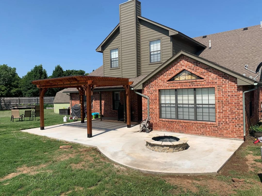 Backyard with brick house, wooden pergola, and stone fire pit under a clear blue sky.